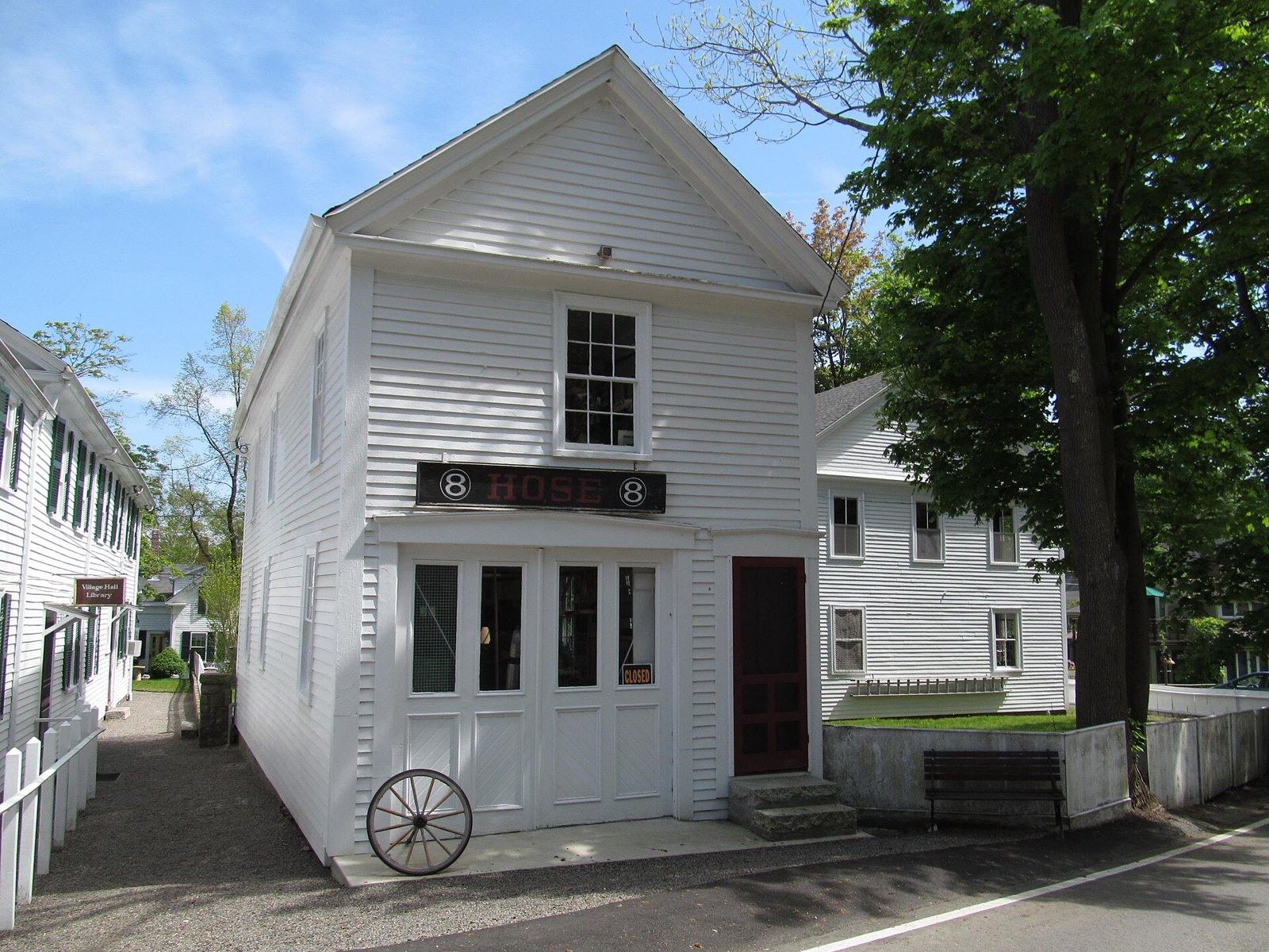 A white clapboard firehouse with a Hose 8 sign over its doors and an old wagon wheel out front, on a tree lined street in Annisquam