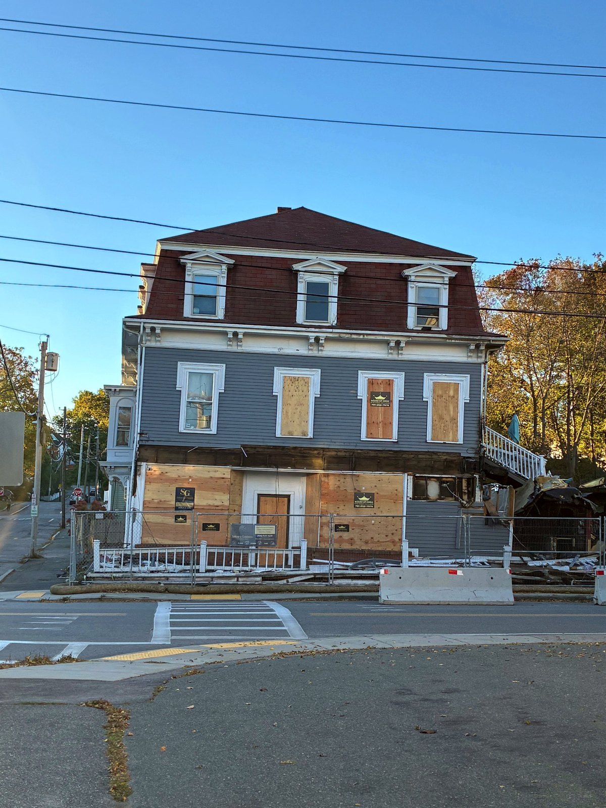 A three story building with a mansard roof on Broadway in Rockport, its first floor and several windows covered with plywood, behind a construction fence with debris piled at one side