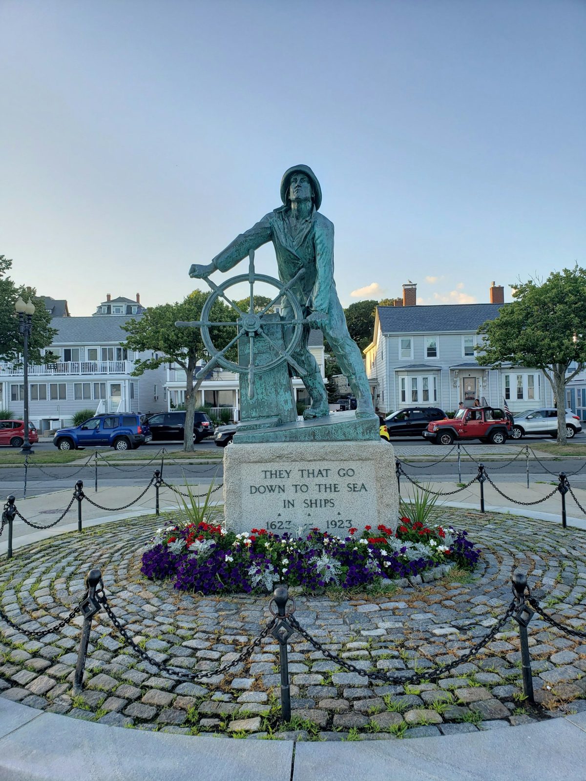 The Gloucester Fisherman's Memorial on Stacy Boulevard: a bronze fisherman at a ship's wheel above an inscribed granite base, with flowers planted around it.
