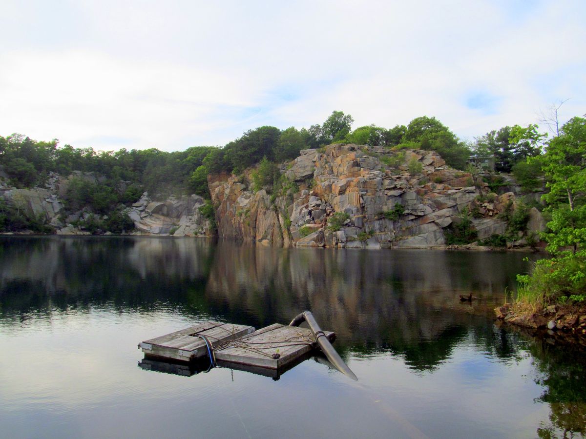 Granite cliffs and trees reflected in Flat Ledge Quarry, with a floating pump intake in the foreground